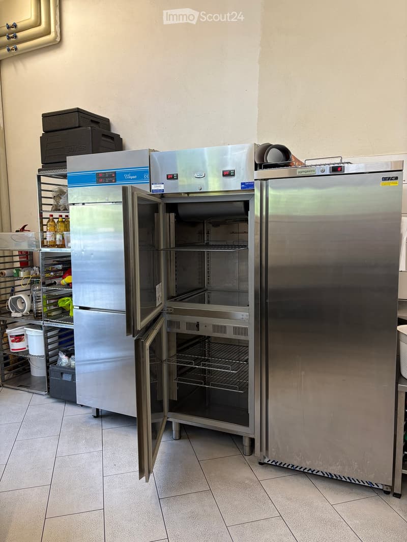 Commercial kitchen with stainless steel refrigerator, oven, and shelving unit holding various items including bottles.