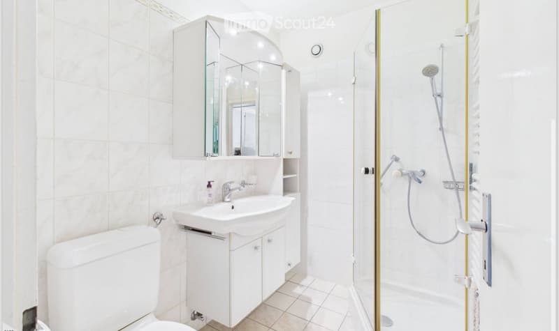 Modern bathroom with white tiles, a toilet, a sink with a mirror cabinet, and a shower with a glass enclosure.