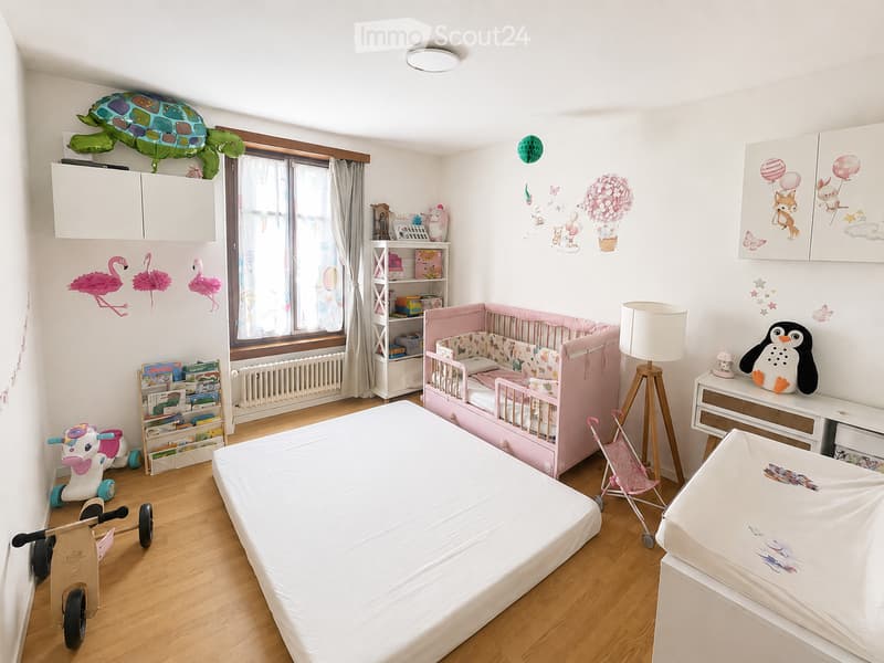 Bright, child-friendly bedroom with a large bed, crib, toy stroller, and various toys. Features wall decals, a bookshelf, and a lamp.