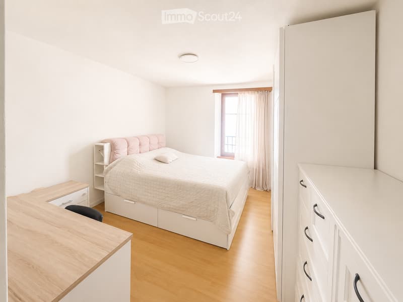 Modern bedroom with a white bed, pink headboard, nightstand, chair, and built-in cabinet. Light wooden flooring and white walls.