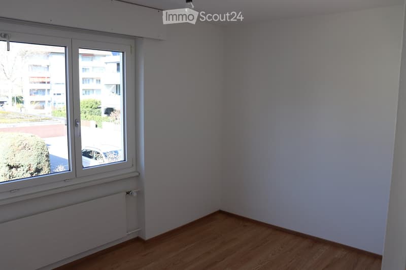 Empty room with wooden flooring, white walls, and a large window with a view of the street and neighboring buildings.