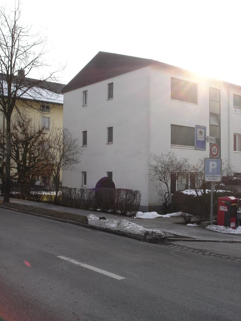 A two-story white building with multiple windows, surrounded by bare trees and bushes, with a parking sign and speed limit sign visible nearby.