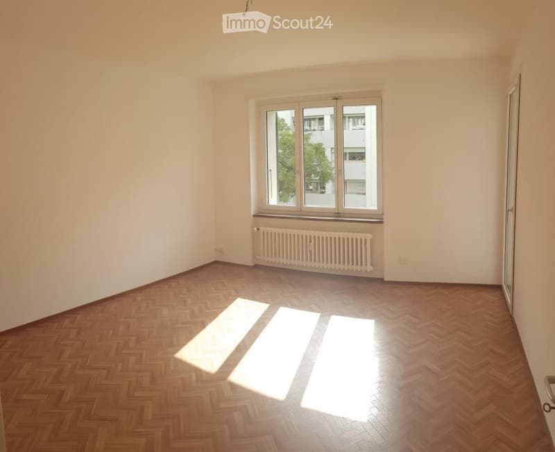Empty room with wooden herringbone flooring, white walls, and a large window with a radiator below it.