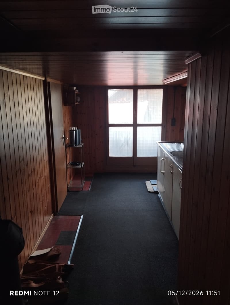 A narrow hallway with wooden panel walls, carpeted floor, and a large window at the end. There are small shelves and a small table with items on the left side.
