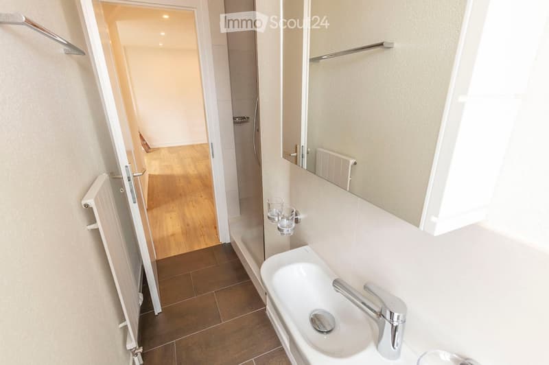 Modern bathroom with a white sink, chrome faucet, large mirror, radiator, and two cups on the wall. The floor is tiled, and the walls are painted in a light color. An open doorway leads to another room with wooden flooring.