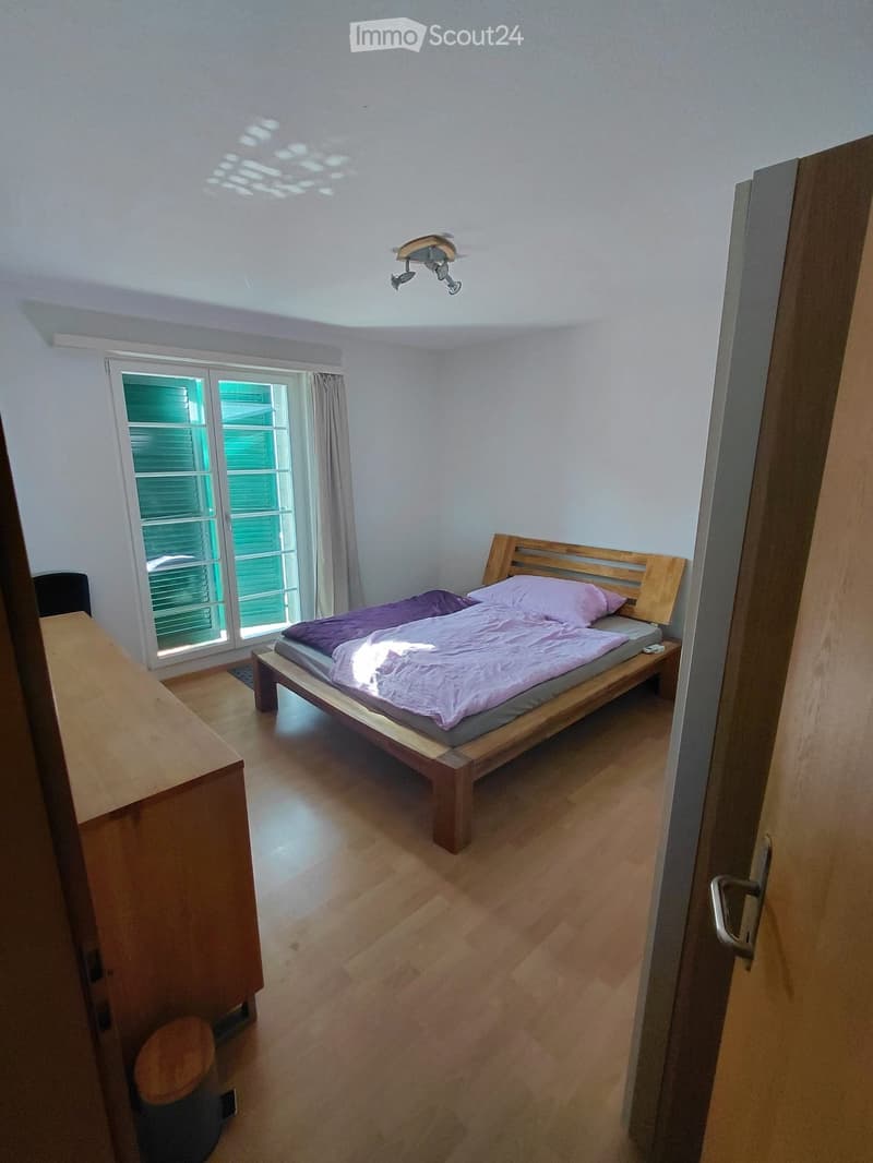 A modern bedroom with a wooden bed, purple bedding, a nightstand, and a small wooden stool. The room features wooden flooring, a large window with blinds, and a ceiling light fixture.