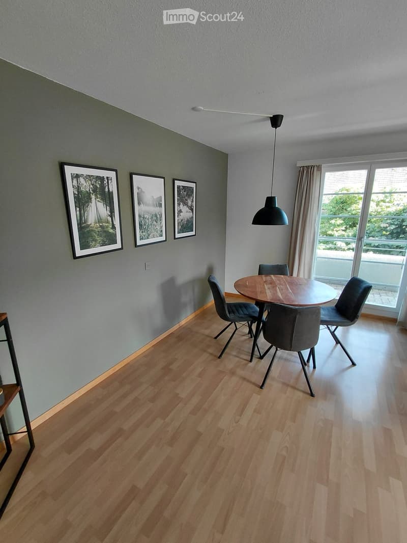 Dining room with wooden floor, round wooden dining table, four chairs, pendant light, and three picture frames on the wall.