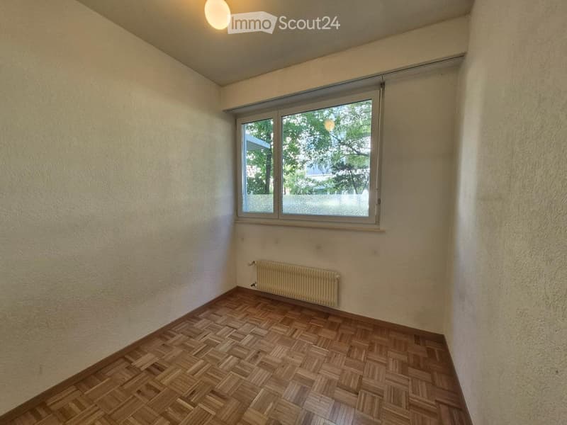 Empty room with wooden parquet flooring, beige walls, a single window with a view of trees, and a radiator below the window.