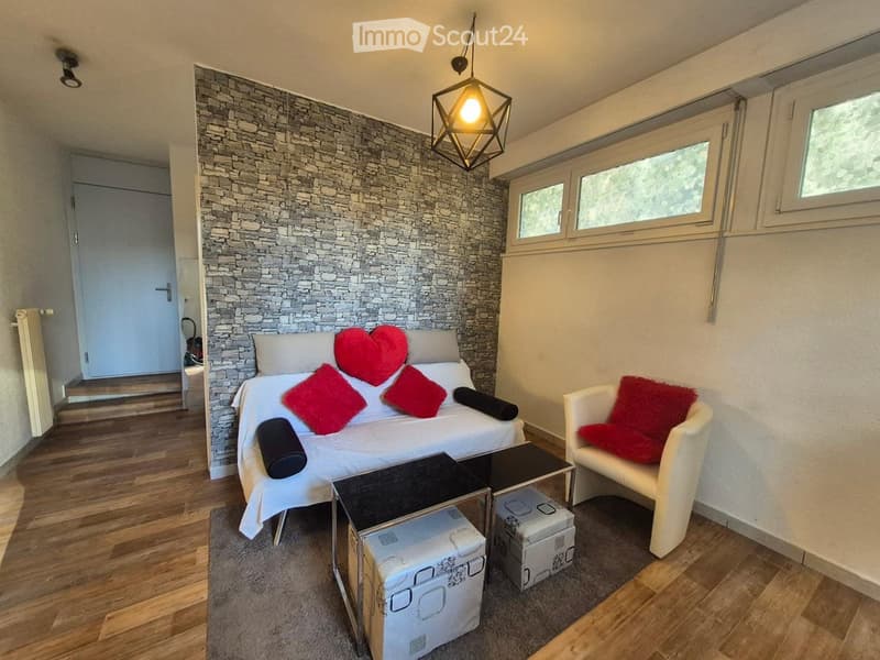 Cozy bedroom with a stone accent wall, a white sofa with red pillows, a white armchair, a small black table, and a pendant light fixture.