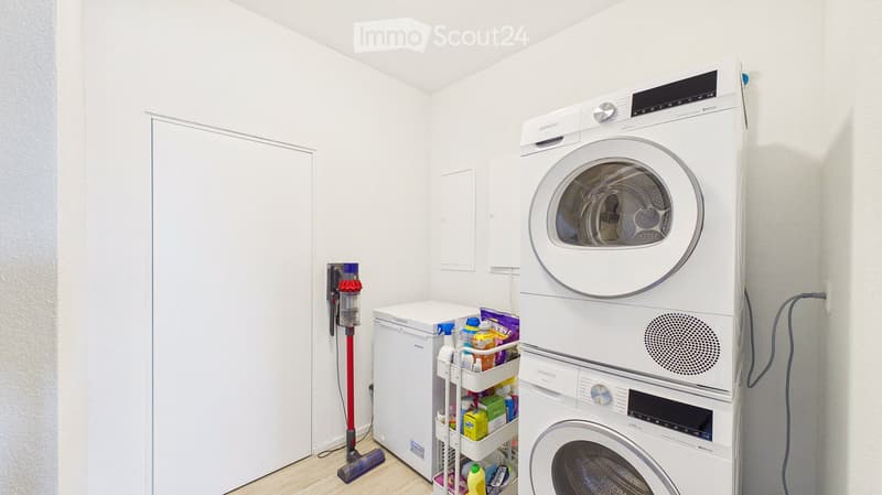 A modern bathroom with a stacked washing machine and dryer, a small refrigerator, cleaning supplies on a cart, and a vacuum cleaner leaning against the wall.