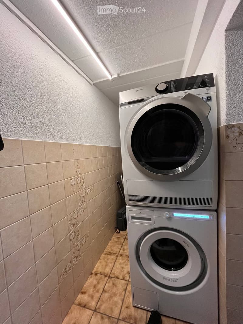 A compact bathroom with a stacked washing machine and dryer unit, beige tiled walls, and a tiled floor.