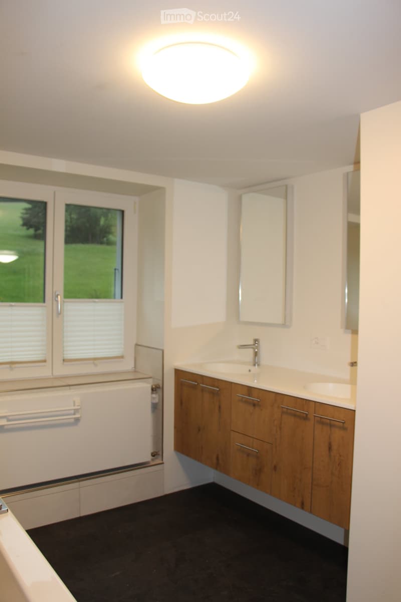 Modern bathroom with a double sink vanity, wooden cabinets, a large mirror, and a bathtub with a window overlooking a green area.