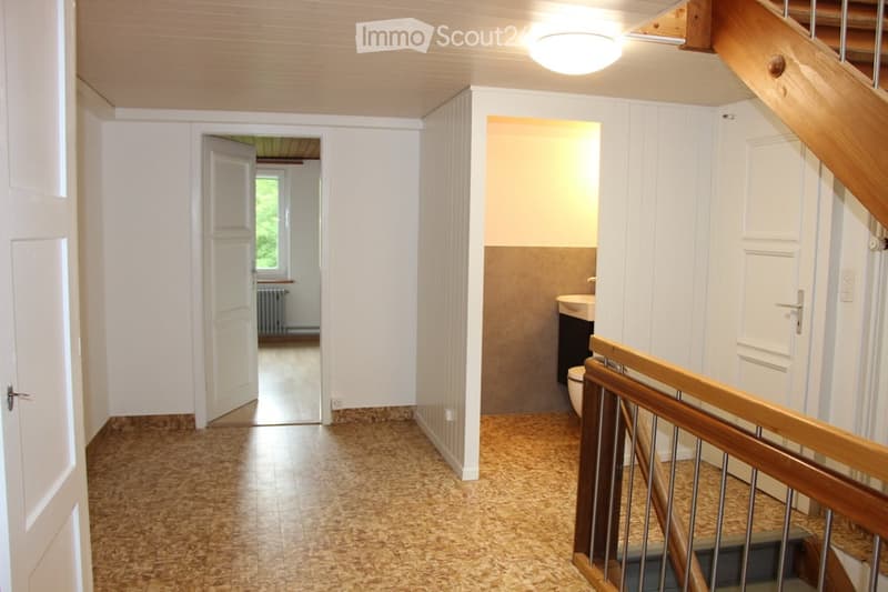 Empty hallway with wooden staircase, white walls, and tiled floor. Doors lead to adjacent rooms, including a bathroom and another room with a window.