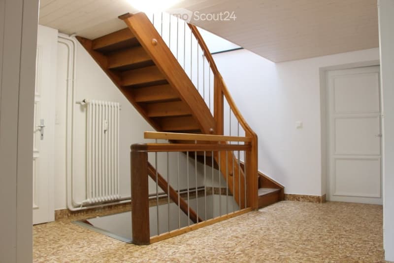 A modern hallway featuring a wooden staircase with metal railings, a radiator on the left wall, and a closed door on the right.