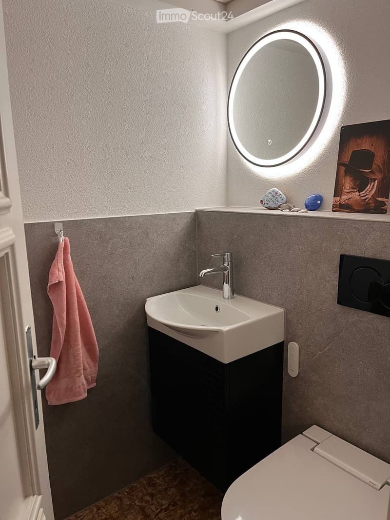 Modern compact bathroom with a white sink, chrome faucet, round illuminated mirror, toilet, and a pink towel hanging on a rack.