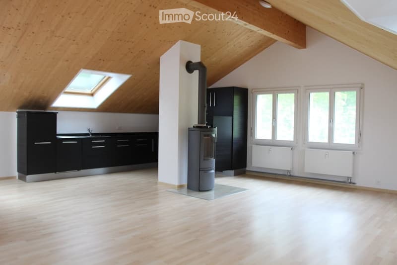 Bright, open room with wooden flooring and exposed wooden beams on the ceiling. Features a modern black kitchen area with cabinets and a sink, a central wood-burning stove with a chimney, and large windows with white frames. The room has a skylight and is