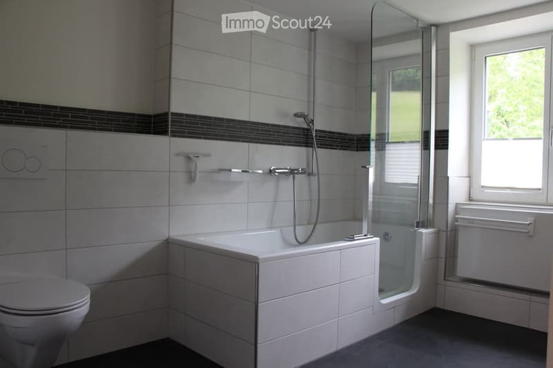 Modern bathroom with a white bathtub, a glass-enclosed shower, a toilet, and a window providing natural light. The walls are tiled with grey tiles and black accents.