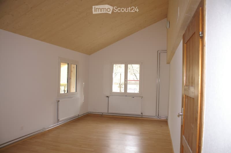 Empty room with wooden flooring, white walls, and exposed wooden ceiling beams. Two windows with white frames and radiators beneath them. A wooden door is visible on the right side.
