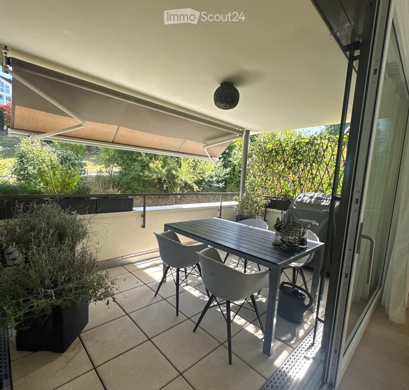 Balcony with a covered area, tiled floor, and a dining set including a table and chairs. There are potted plants and a view of greenery outside.