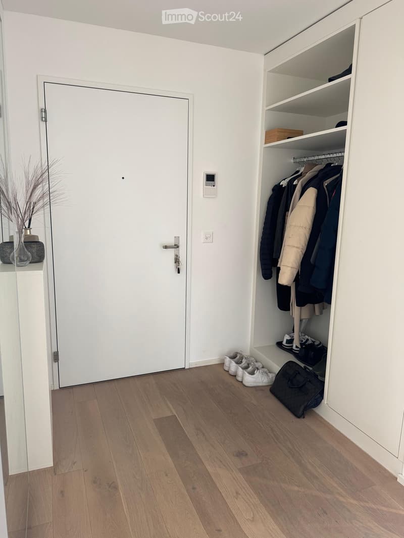 A modern, minimalist hallway with a built-in wardrobe, wooden flooring, and a small table with a vase and decorative items.