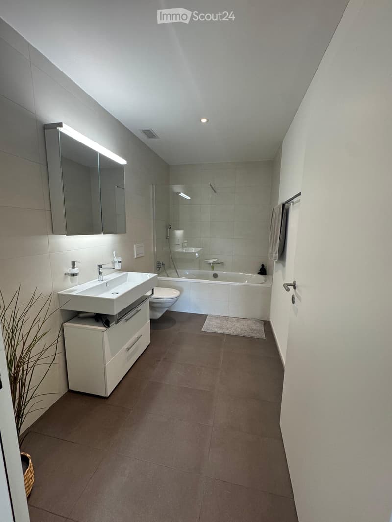 Modern bathroom with a white sink, mirror with built-in lighting, toilet, bathtub, and a small cabinet. The space features light grey tiles and a sleek design.