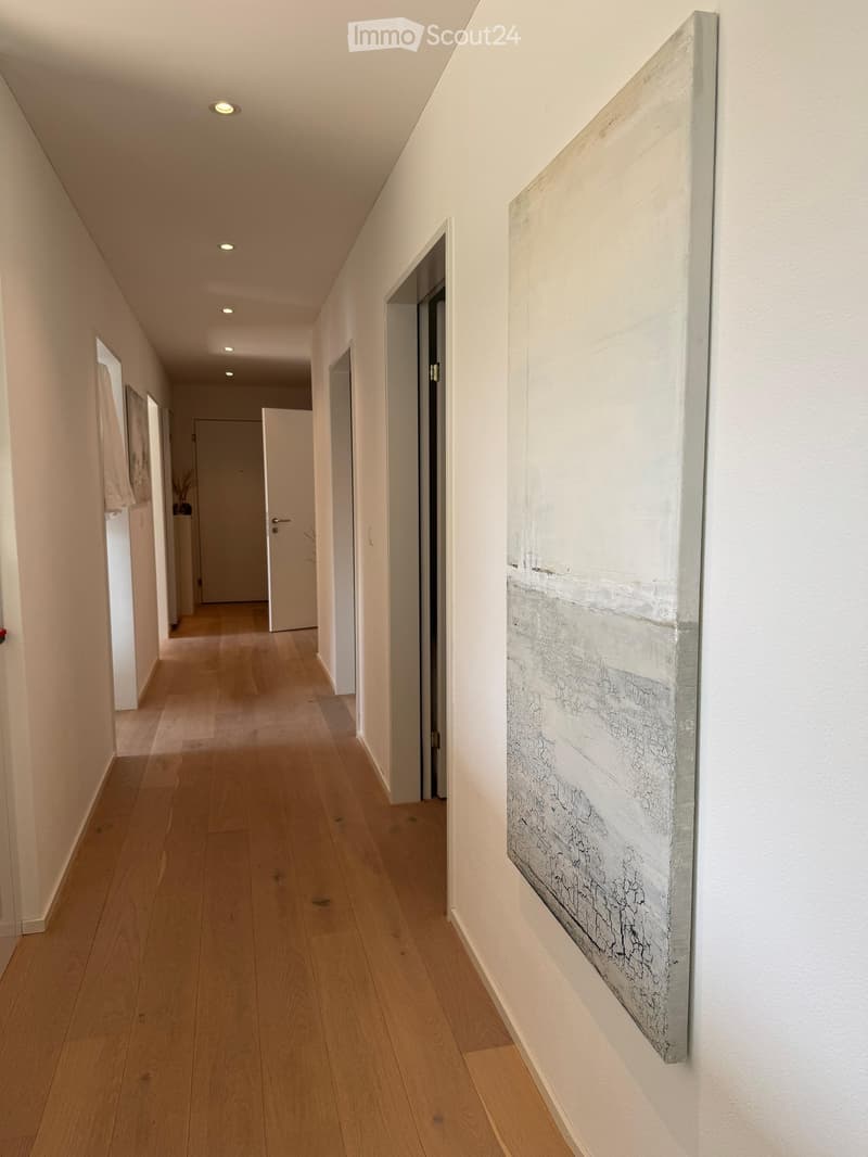 A modern hallway with light wooden flooring, white walls, recessed ceiling lights, and a large abstract painting on the right wall. Doors lead to other rooms.