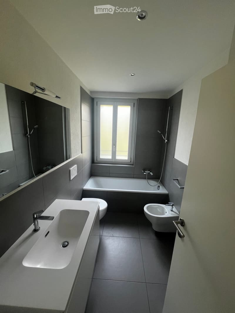 Modern bathroom with grey tiles, a large sink, toilet, bathtub, and a window providing natural light.