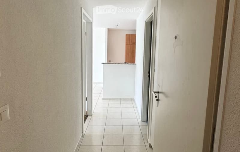 A narrow hallway with tiled flooring, white walls, and a small white counter in the background. There is a wooden cabinet visible in the distance.