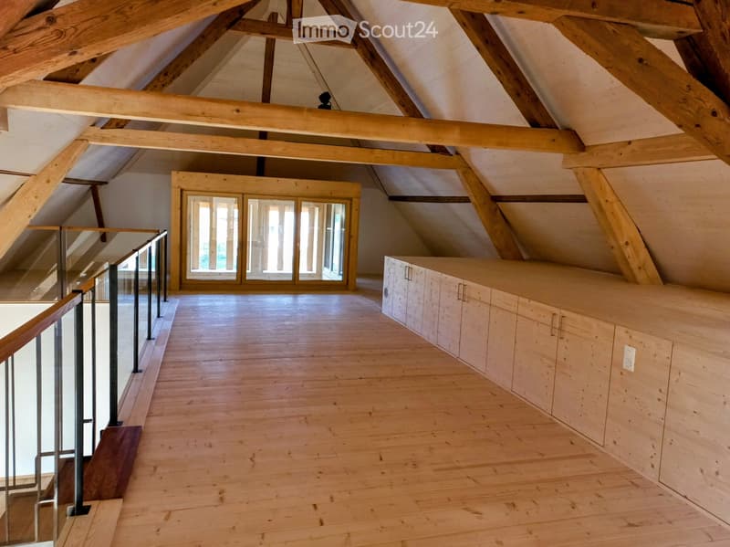 Open, airy attic space with exposed wooden beams, wooden flooring, built-in cabinetry, and large glass doors leading to a balcony.