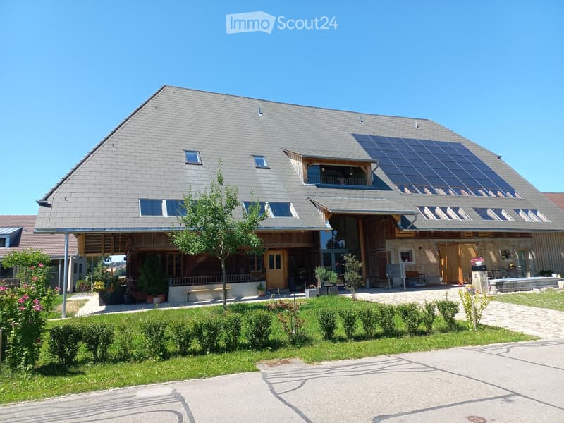 A modern home with a sloping roof, solar panels, glass windows, a balcony with wooden railings, potted plants, a bench, and a green lawn. A pathway leads to the entrance, and there are flowers and shrubs in the foreground.
