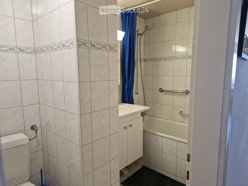 Bathroom with white tiled walls, a toilet, a bathtub with a shower, a sink with a cabinet, and a handrail for safety.