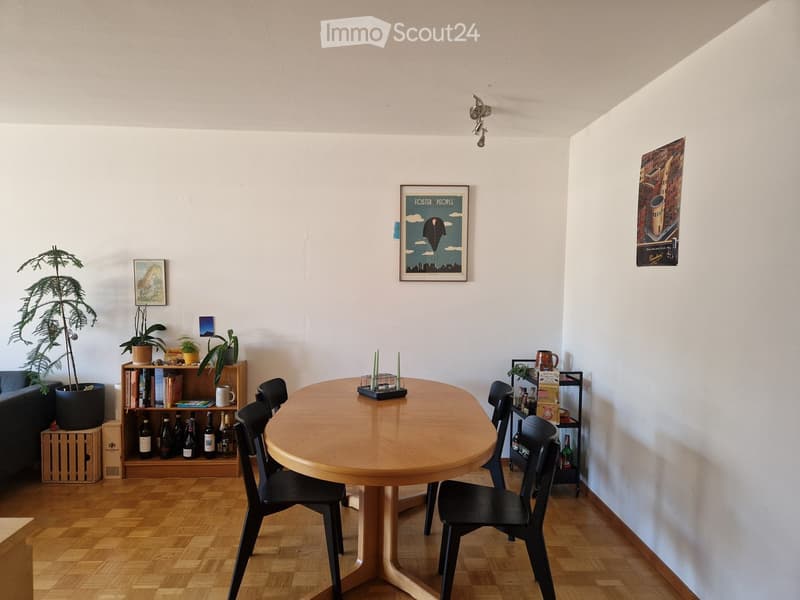 A modern dining area with a round wooden table surrounded by black chairs, shelves with books and bottles, potted plants, and artwork on the walls.