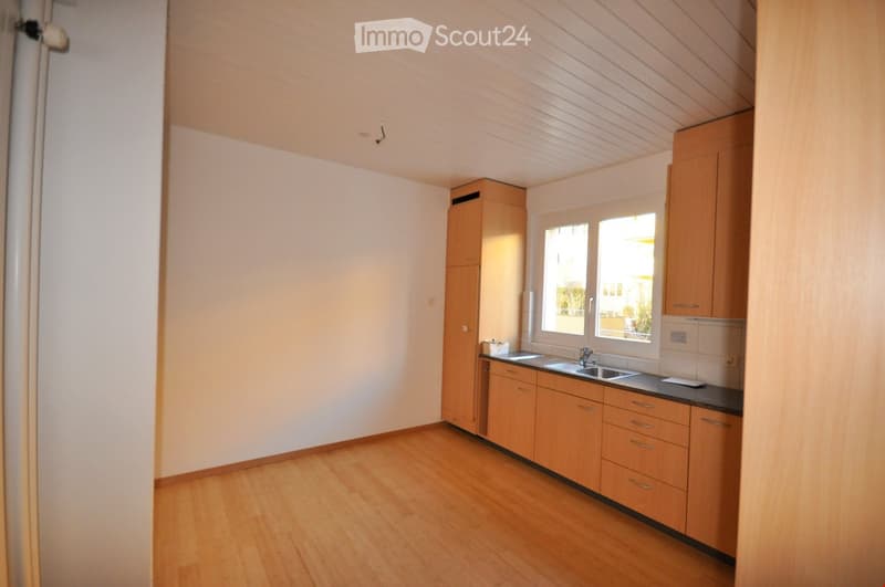 Bright, unfurnished kitchen with light wooden cabinets, a sink, and a countertop. The space features a wooden floor and large windows allowing natural light to enter.