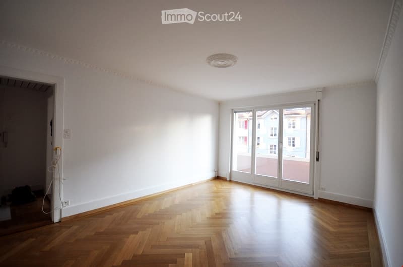 Empty room with wooden parquet flooring, white walls, large sliding glass doors leading to a balcony, and a ceiling light fixture.