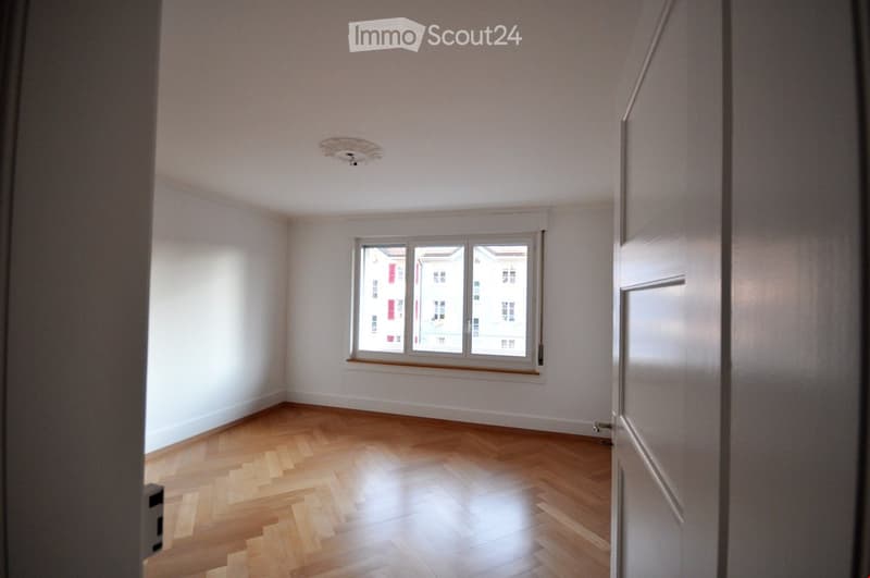 Empty room with light wooden flooring, white walls, and a single window showing a view of neighboring buildings.