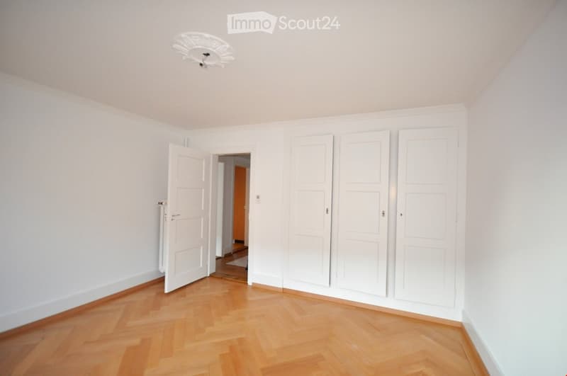 Empty room with light-colored wooden flooring, white walls, and several closed white doors. A ceiling light fixture is visible.