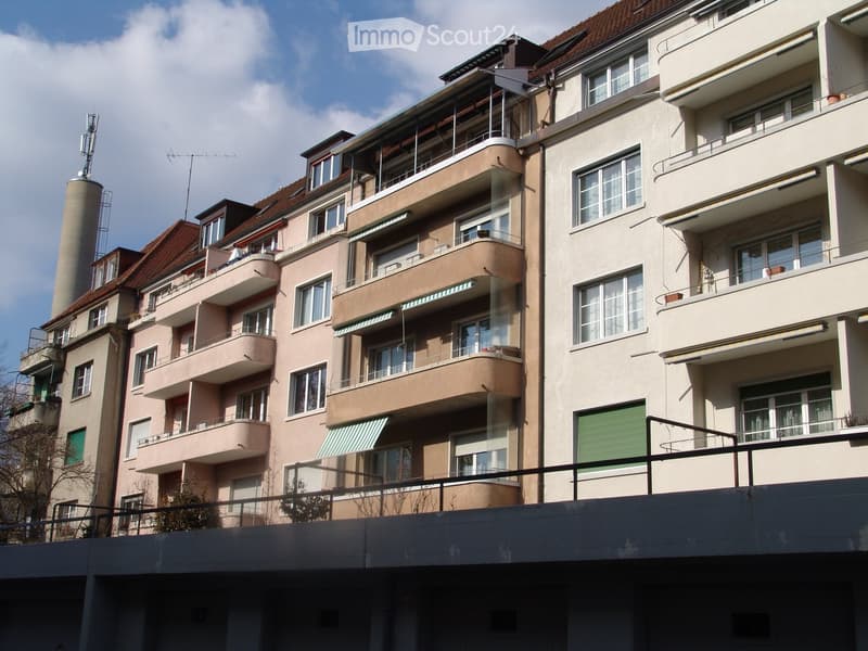 Multi-story residential building with balconies, large windows, and awnings. The structure features a mix of beige and pink facades with green shutters on some windows.