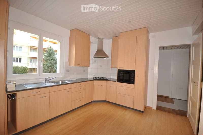 Modern kitchen with light wooden cabinets, stainless steel sink, stove, microwave, and range hood. Large windows provide natural light.