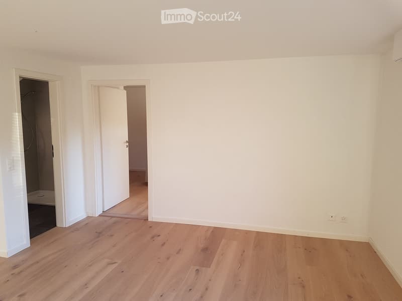 Empty room with light wooden flooring and white walls, featuring two open doors leading to other spaces.