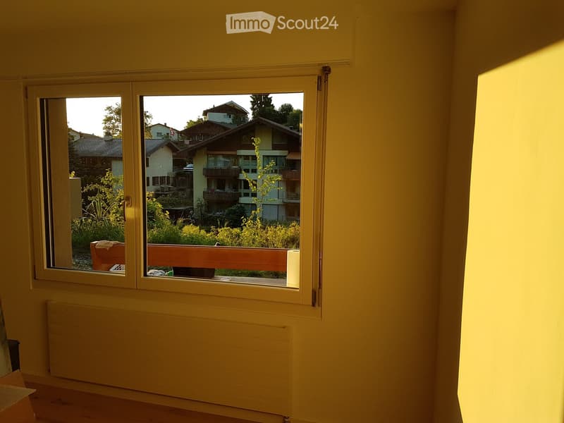 Empty room with light yellow walls, wooden floor, and a large window offering a view of a residential area with houses and greenery.
