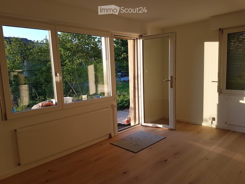 Empty room with wooden flooring, large windows, and a sliding glass door leading to a garden area.