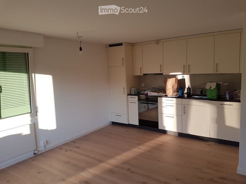 Bright, modern kitchen with white cabinets, wooden flooring, and a window with blinds. Visible items include an oven, sink, and various kitchen items on the counter.
