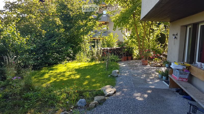 A well-maintained garden with a gravel pathway, lush greenery, potted plants, and a wooden bench near the house.