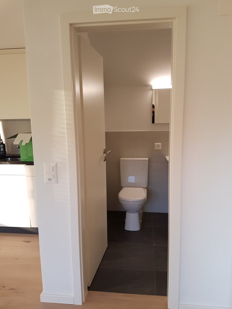 Small bathroom with a toilet, dark tiled floor, and white walls. The door is open, revealing the space.