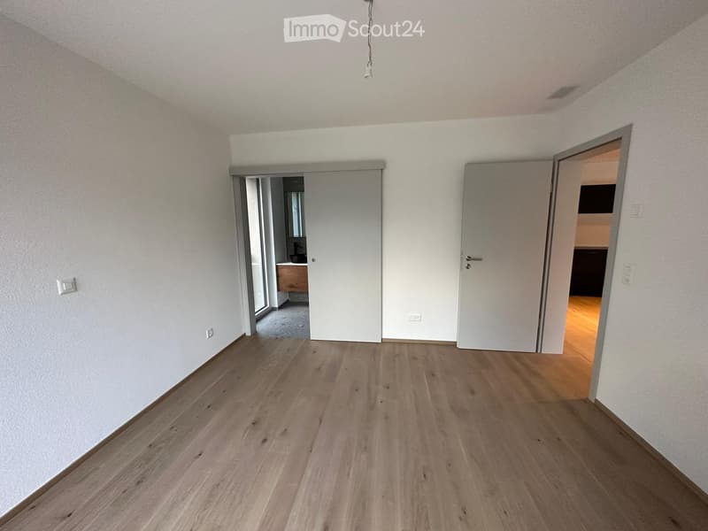 Empty room with light wooden flooring and white walls, featuring two open doors leading to other rooms.