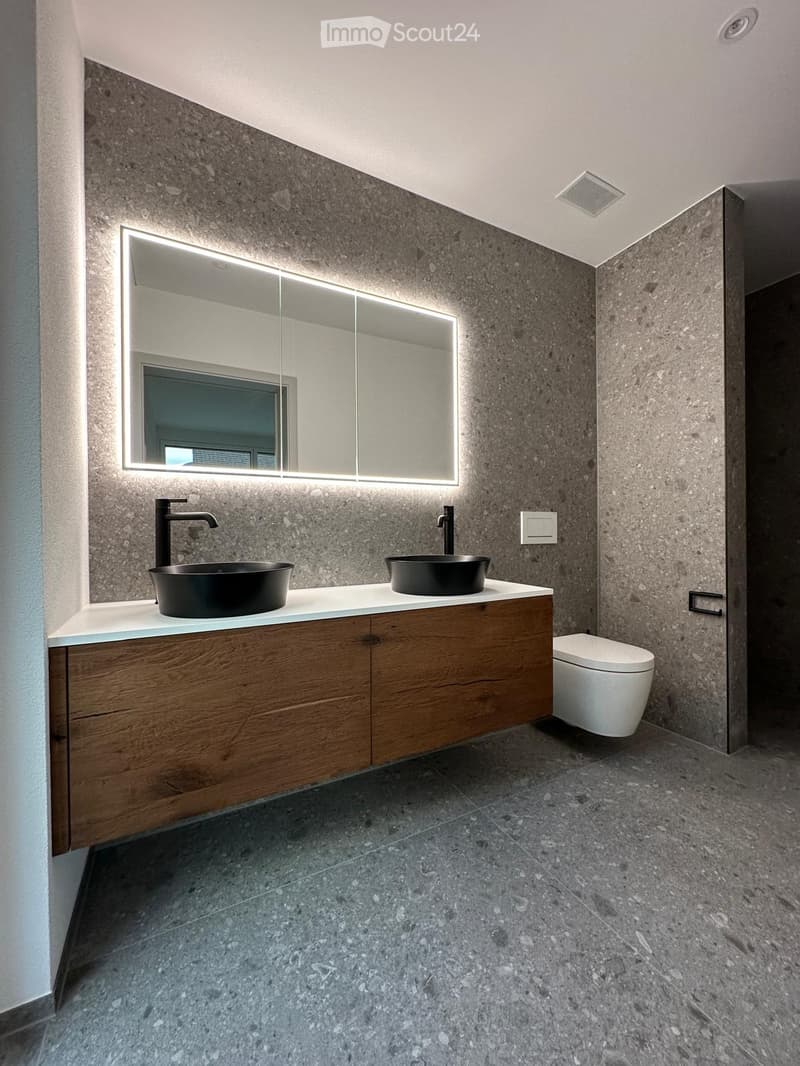 Modern bathroom with dark stone walls, two black sinks, wooden cabinet, large mirror with LED lighting, and a toilet.