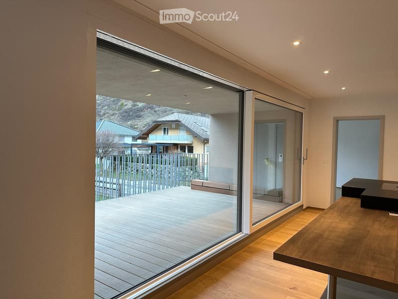 Modern interior with large glass sliding doors leading to a wooden deck, a wooden table, and a view of the outdoor area and neighboring houses.