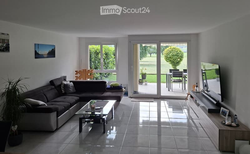Modern living room with a dark sectional sofa, glass coffee table, TV screen, and large windows overlooking a green garden. The room features a tiled floor and a sliding glass door leading to a patio.