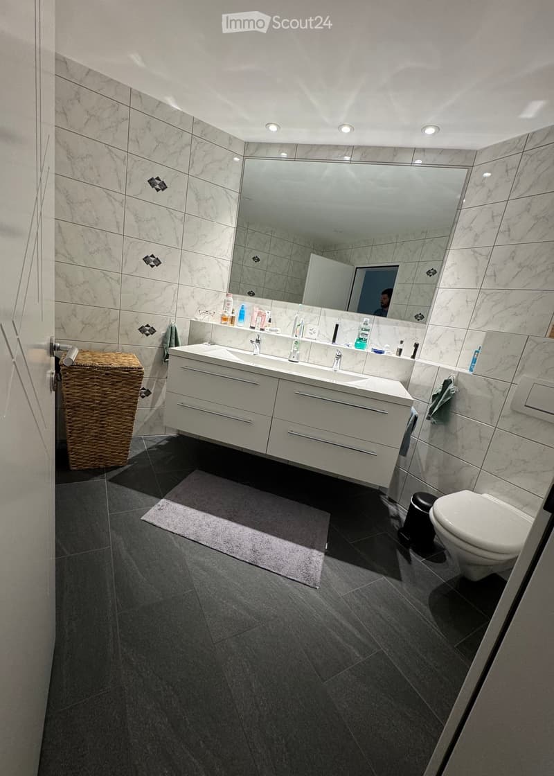 Modern bathroom with marble tiles, double sink vanity, large mirror, toilet, wicker basket, and a small rug on the floor.