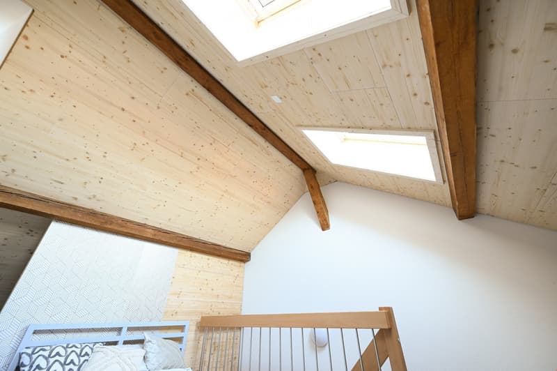 A modern, minimalist bedroom with wooden flooring, white walls, and a slanted wooden ceiling with skylights. A bed with patterned pillows is visible in the foreground.